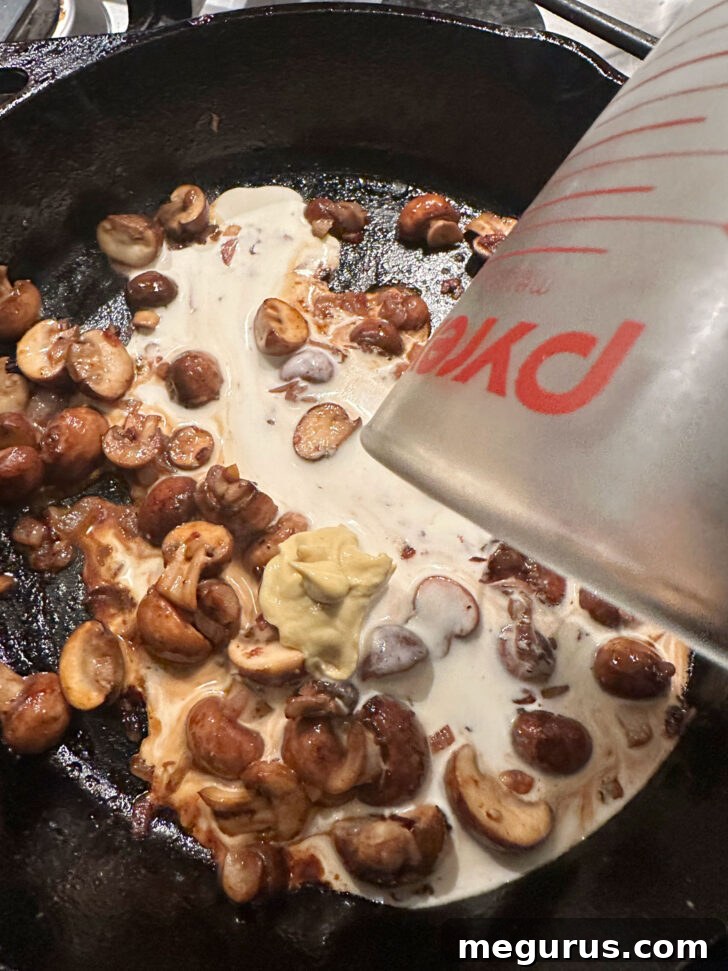 Heavy cream and Dijon mustard being stirred into the mushroom mixture in the skillet, forming the creamy base of the sauce.