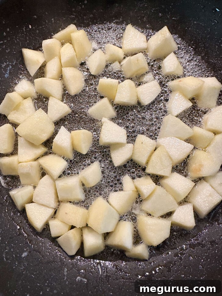 Diced tart apples cooking in a skillet with a small amount of butter, softening and releasing their liquid.