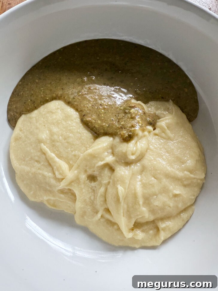 The smooth, green pistachio mixture being added to the reserved plain cake batter in a bowl.
