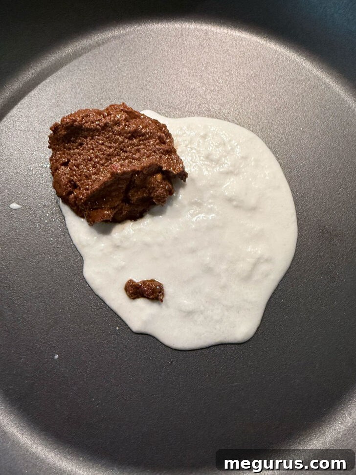 Thick coconut cream being heated with vibrant curry paste in a skillet.