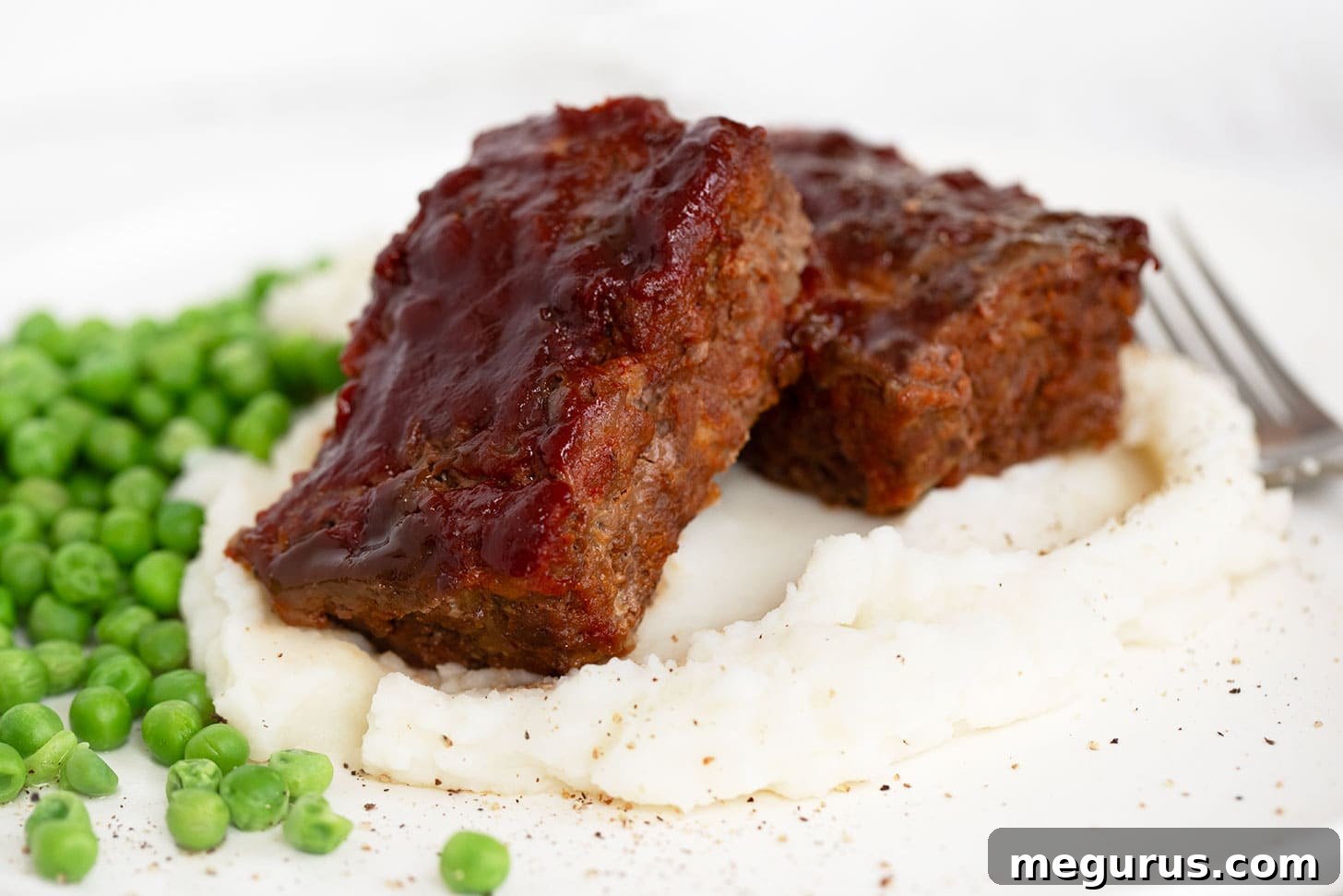 Flavorful Meatloaf with homemade BBQ sauce, served with creamy mashed potatoes and vibrant green peas.