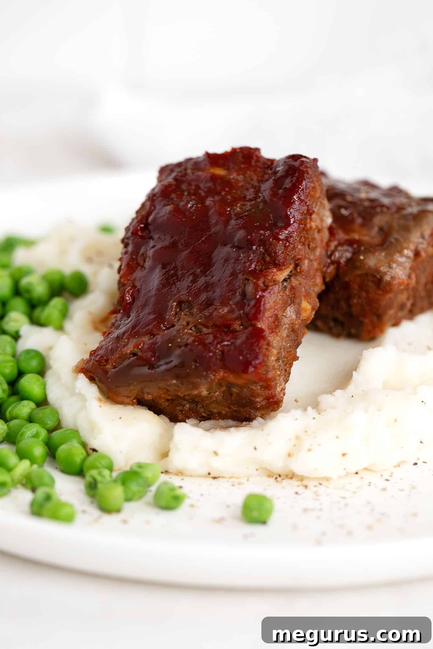 A serving of homemade BBQ meatloaf with creamy mashed potatoes and fresh green peas on a dinner plate, ready to enjoy.