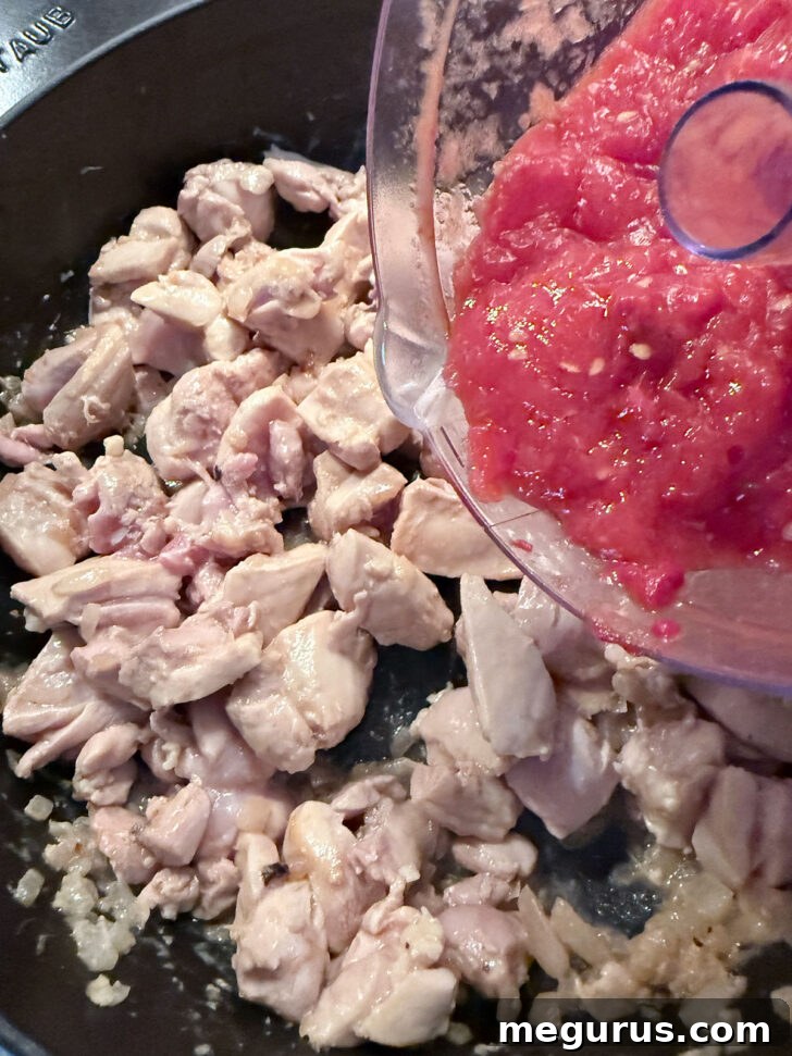 Smooth pureed tomatoes being stirred into the skillet with the cooked chicken and onion mixture, creating the rich curry foundation.