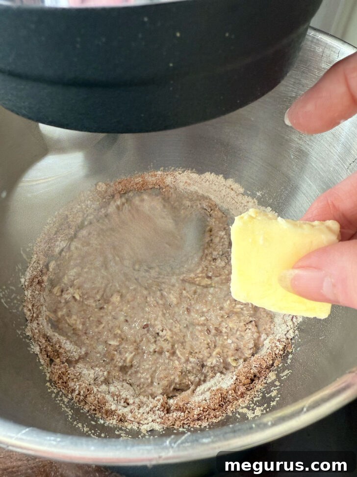 Adding softened butter to the mixing bowl with flour and grains.