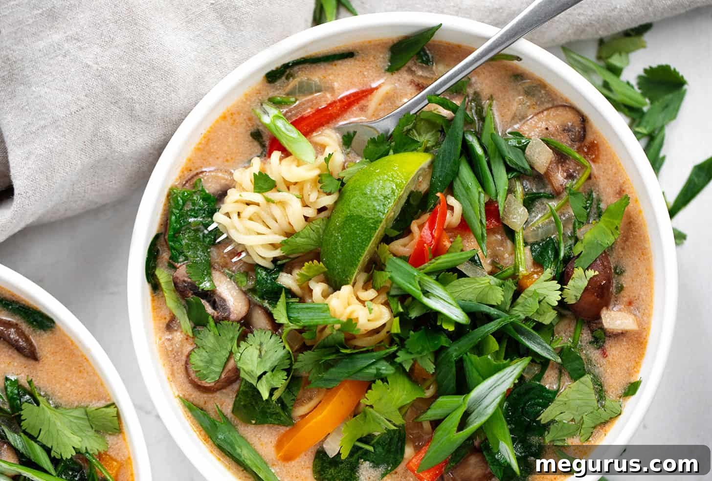 A close-up of a bowl of spicy coconut ramen, garnished with fresh herbs and a fork ready to pick up noodles.