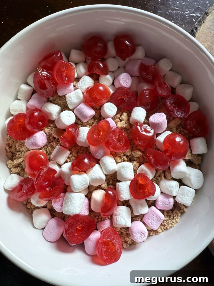 Crushed Digestive biscuits, brightly colored glacé cherries, and soft marshmallows mixed together in a large mixing bowl, awaiting the binding agent.