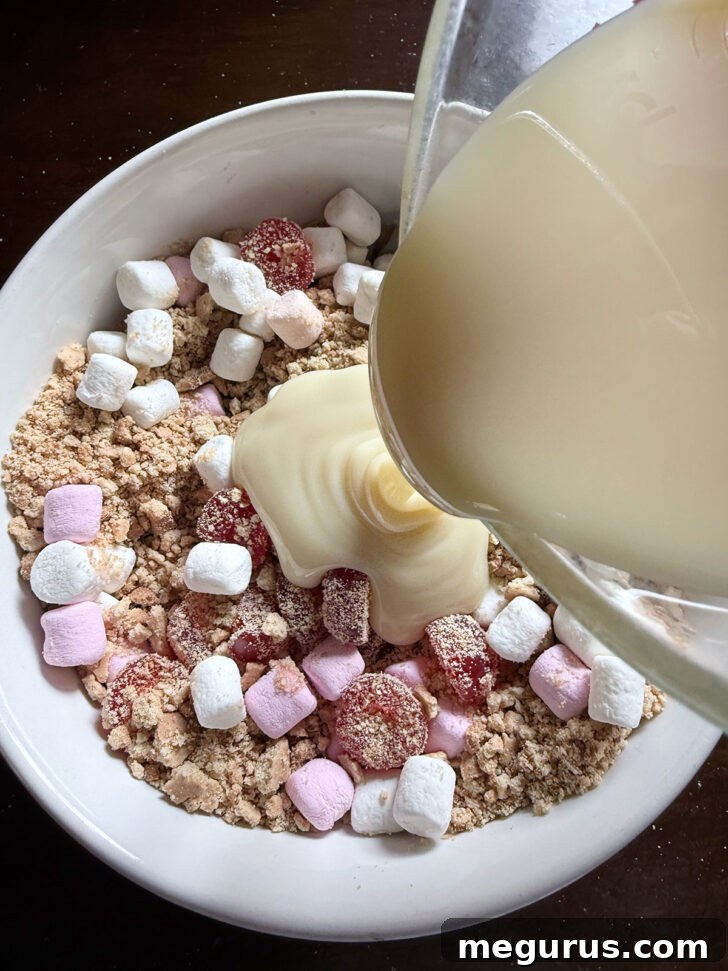 Sweetened condensed milk being poured into the bowl containing the biscuit, marshmallow, and cherry mixture, initiating the binding process.