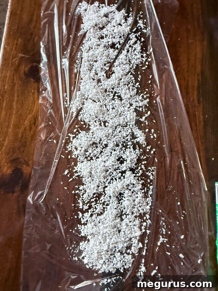 A long piece of plastic wrap spread on a counter, with a generous line of desiccated coconut sprinkled down the middle, preparing for the mixture.