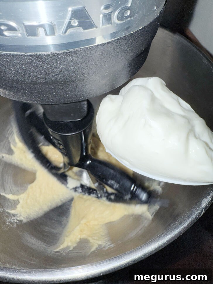 A view of sour cream being carefully added to the coffee cake batter in a mixer, highlighting the alternate mixing technique.