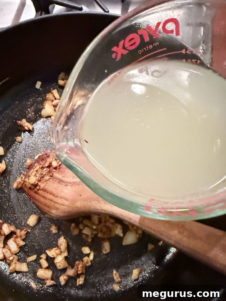 Pouring chicken broth into the skillet with softened onions and curry paste.