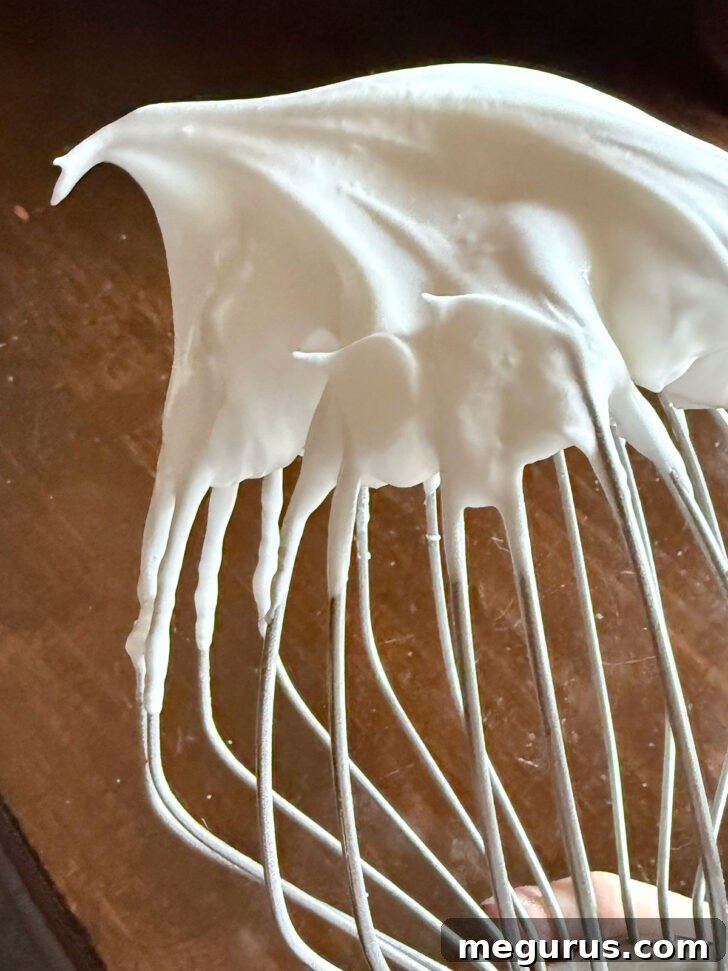 Stiffly whipped egg whites clinging to the whisk attachment of a stand mixer, indicating perfect meringue consistency.