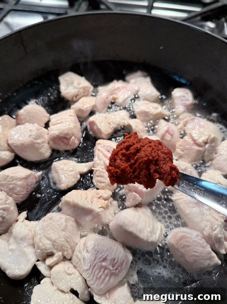Adding fragrant Thai red curry paste to the beautifully browned chicken pieces in a hot skillet, ensuring even coating.