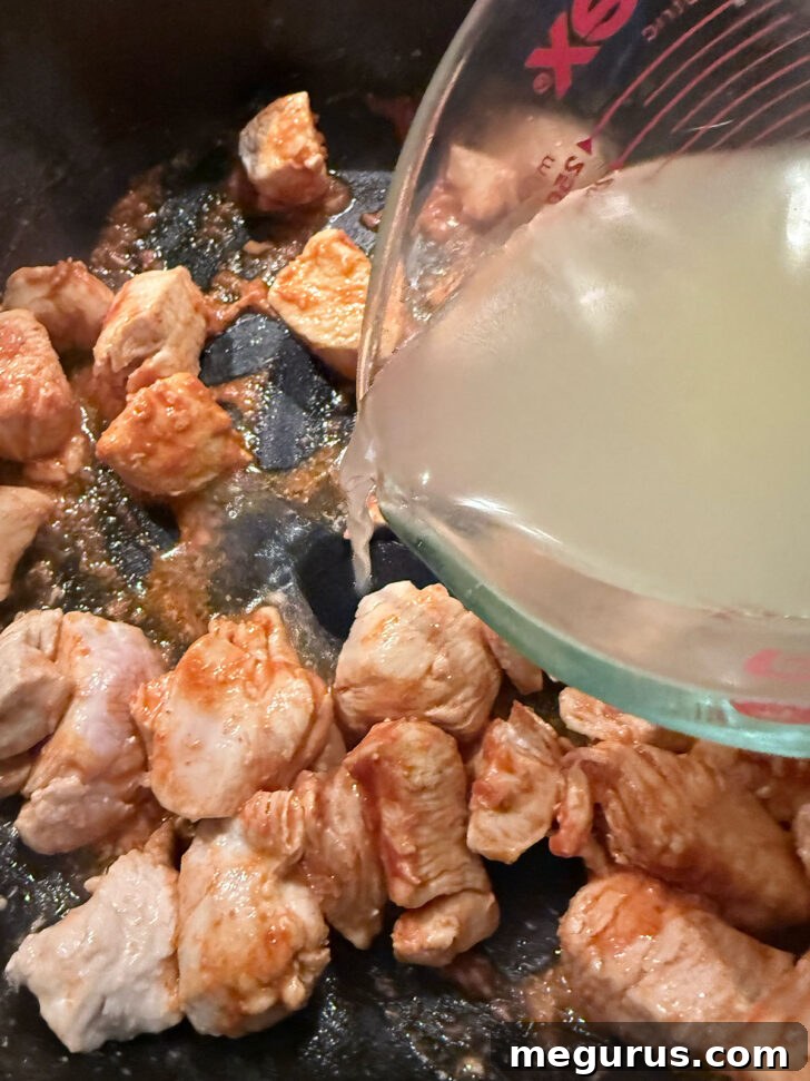 Pouring rich chicken broth into the skillet with chicken and curry paste, preparing for the vegetables.