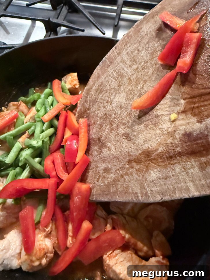 Adding vibrant bell pepper and fresh green beans to the simmering chicken and curry mixture in the skillet.