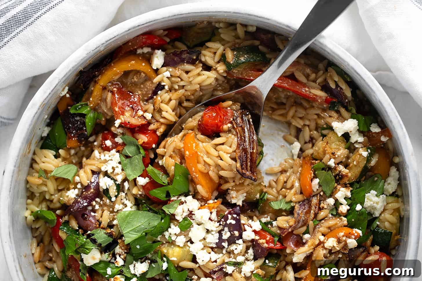 Roasted vegetable orzo in a serving bowl with a spoon, garnished with fresh basil.
