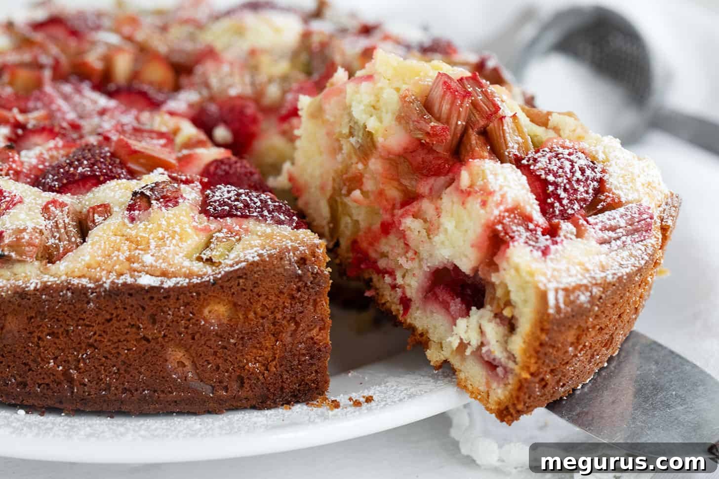 A slice of homemade strawberry rhubarb cake on a white serving plate, showing its moist texture and abundant fruit.