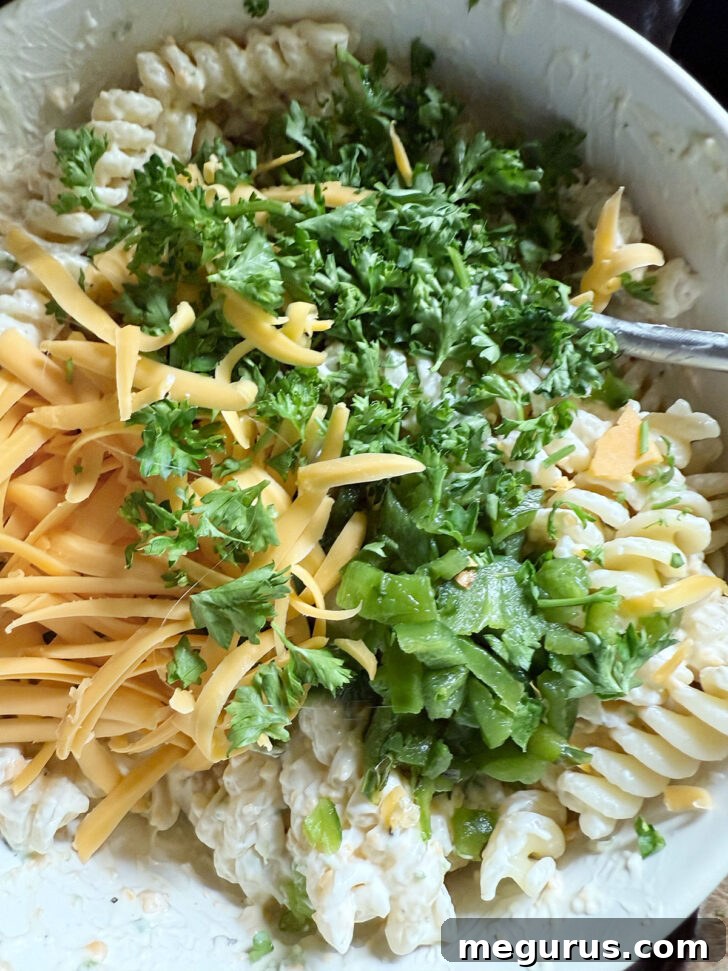 Chopped roasted jalapeño, fresh chopped parsley, and shredded cheddar cheese added to the pasta and dressing in a bowl, ready to be tossed.