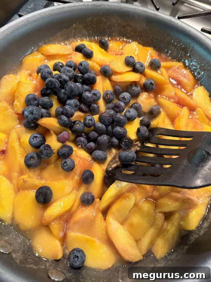Fresh blueberries being added to the simmering peach mixture in the skillet.