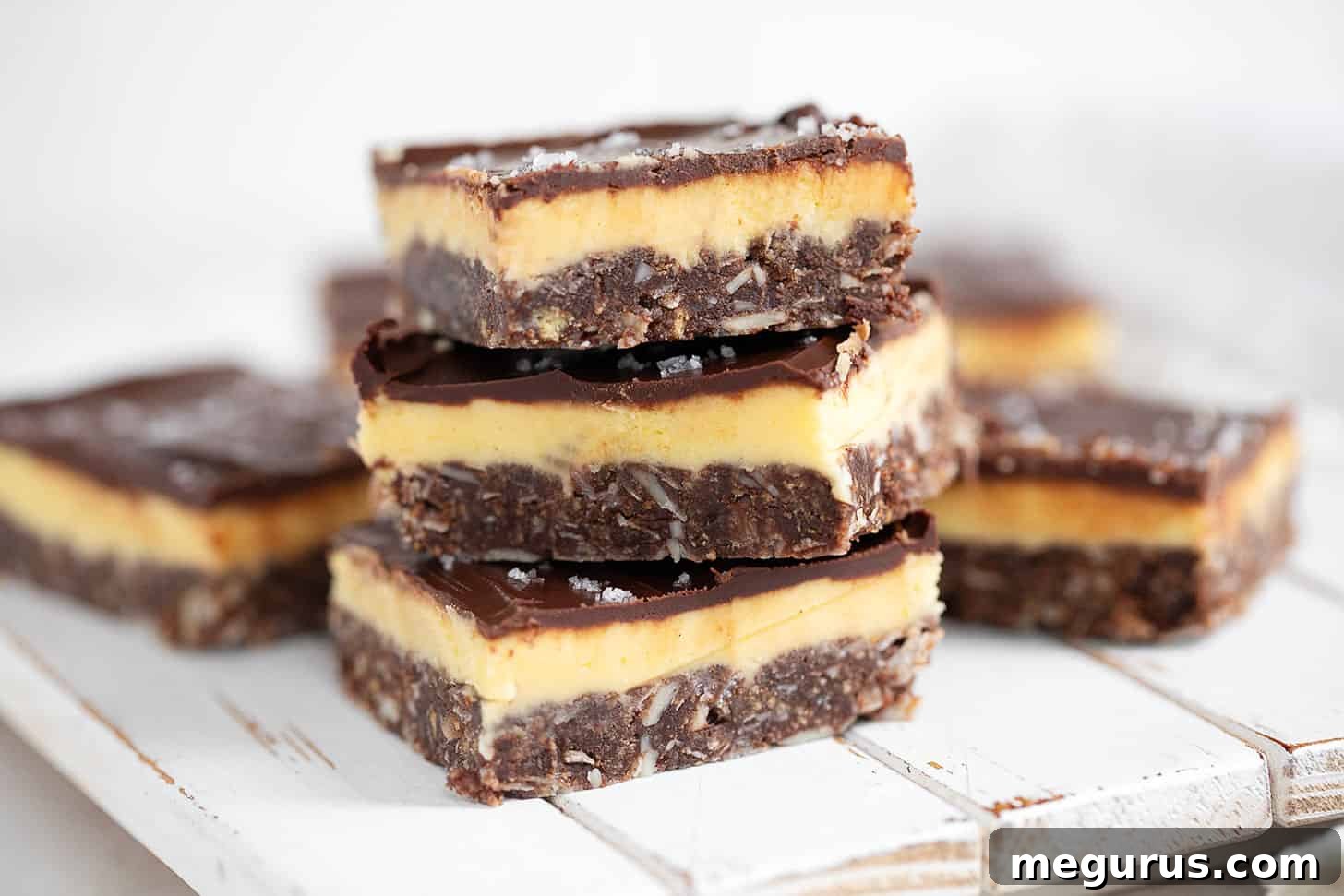 Nanaimo bars cut into perfect squares and stacked elegantly, showcasing their distinct three layers: a dark chocolate coconut base, a creamy yellow custard filling, and a smooth chocolate topping.