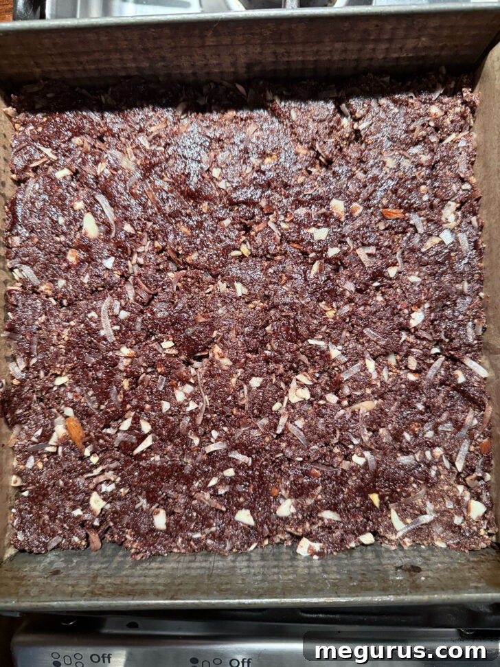 The dark chocolate base layer of Nanaimo bars firmly pressed into a parchment-lined 8x8 inch baking pan, ready for the next layer.