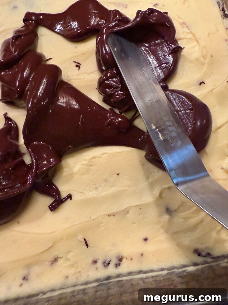 Melted chocolate and butter being poured and spread smoothly over the creamy custard filling layer in the pan, forming the top layer.