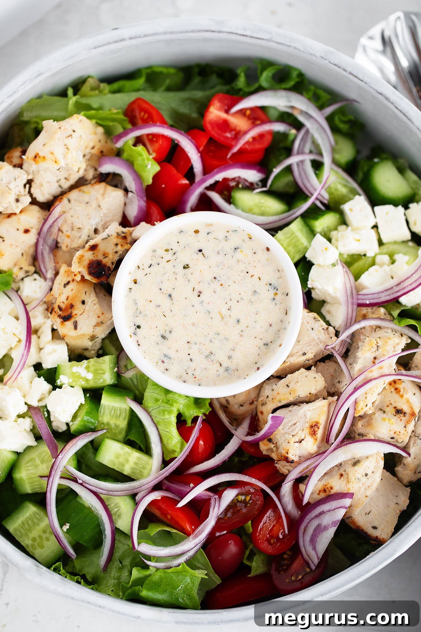 Grilled Chicken Souvlaki Salad with Tzatziki and Lemon Dressing 7 A close-up of a chicken souvlaki salad featuring grilled chicken, cherry tomatoes, cucumber, red onion, lettuce, feta cheese, and a small bowl of creamy dressing in the centre.